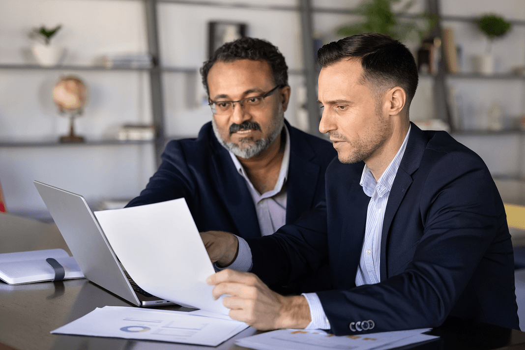 Two colleagues in navy blazers review printed financial reports beside an open laptop at an office table.