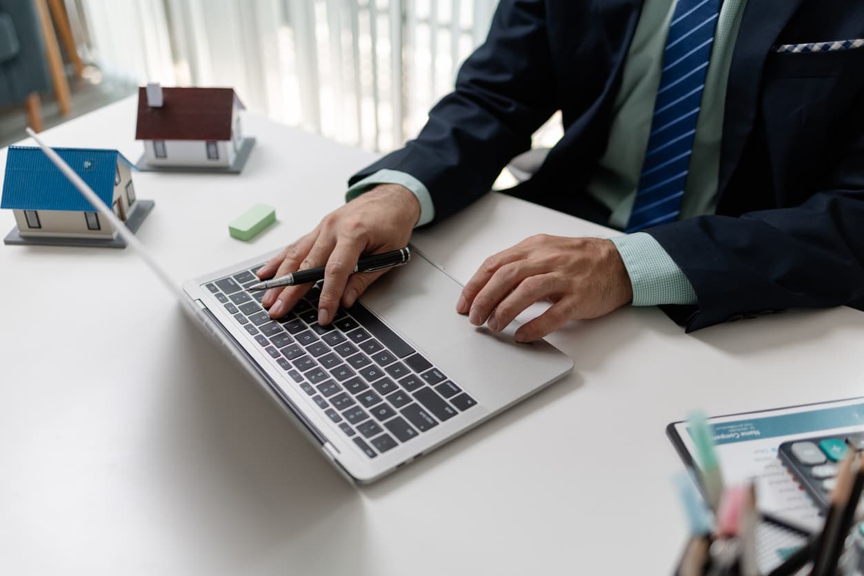 A property manager working at a laptop with model houses and certification paperwork on the desk.