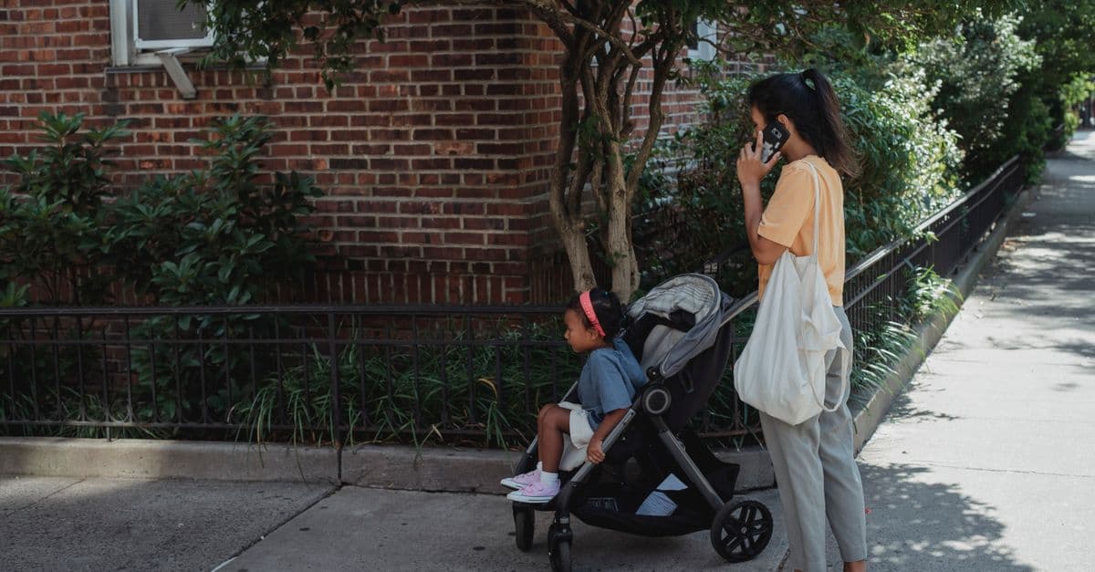 A person on a phone call on the sidewalk outside a brick apartment building, with a child in a stroller nearby.