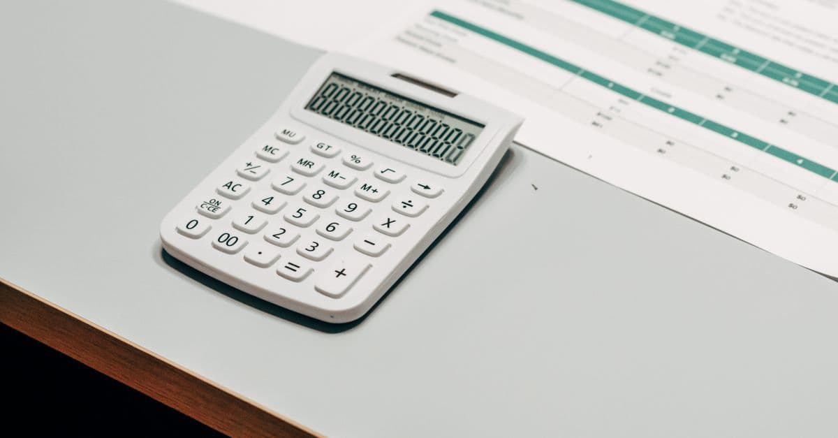 A white desk calculator sitting beside a printed financial spreadsheet on a gray desk.