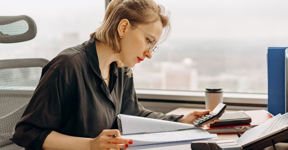 A compliance staffer at a desk running numbers on a calculator against a stack of paperwork, the kind of hand-checked math behind a Total Tenant Payment calculation.