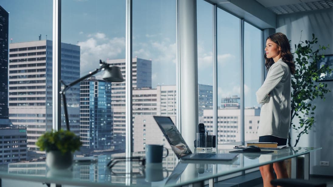 A property executive in a white blazer stands at a floor-to-ceiling office window, arms folded, looking out over a city skyline, with a laptop open on a glass desk behind her.