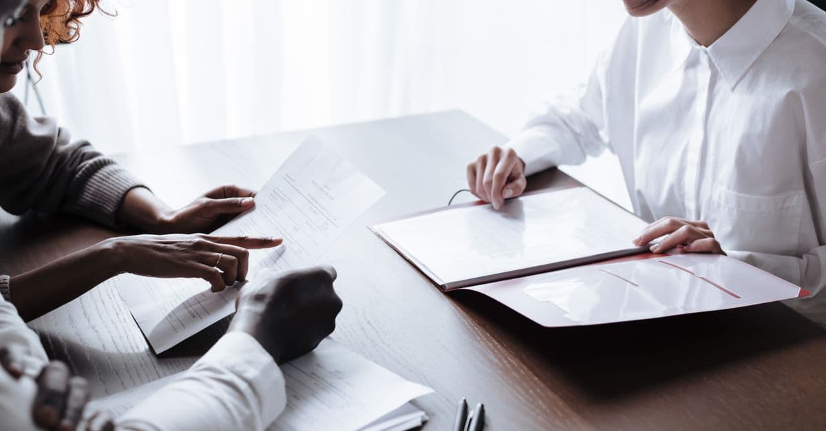 Three people at a desk reviewing printed forms together, one pointing to a line on a page while another holds an open folder of documents.