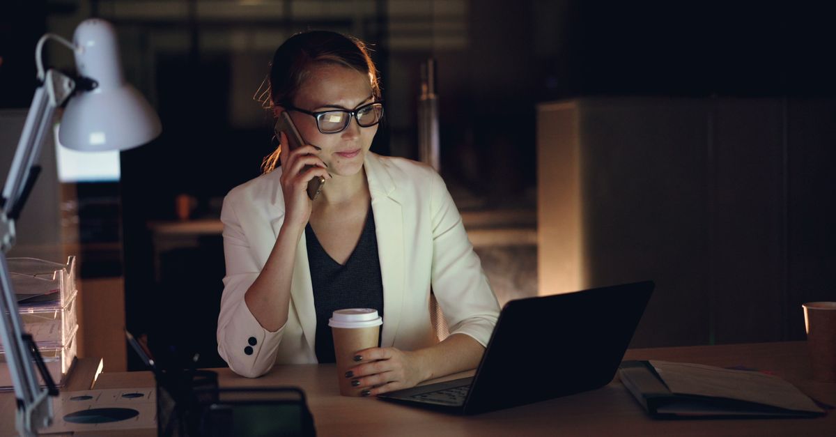 A leasing manager works late at a laptop, holding coffee and taking a call.