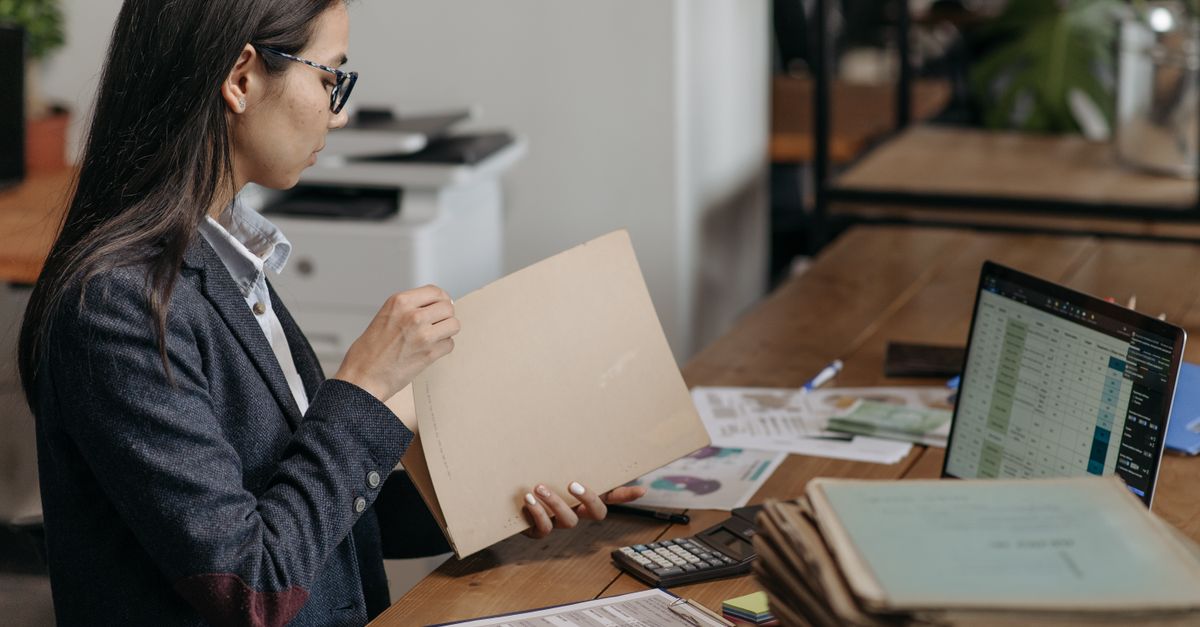 An office worker checks a paper file folder at a desk stacked with binders, a calculator, and a laptop showing a spreadsheet.