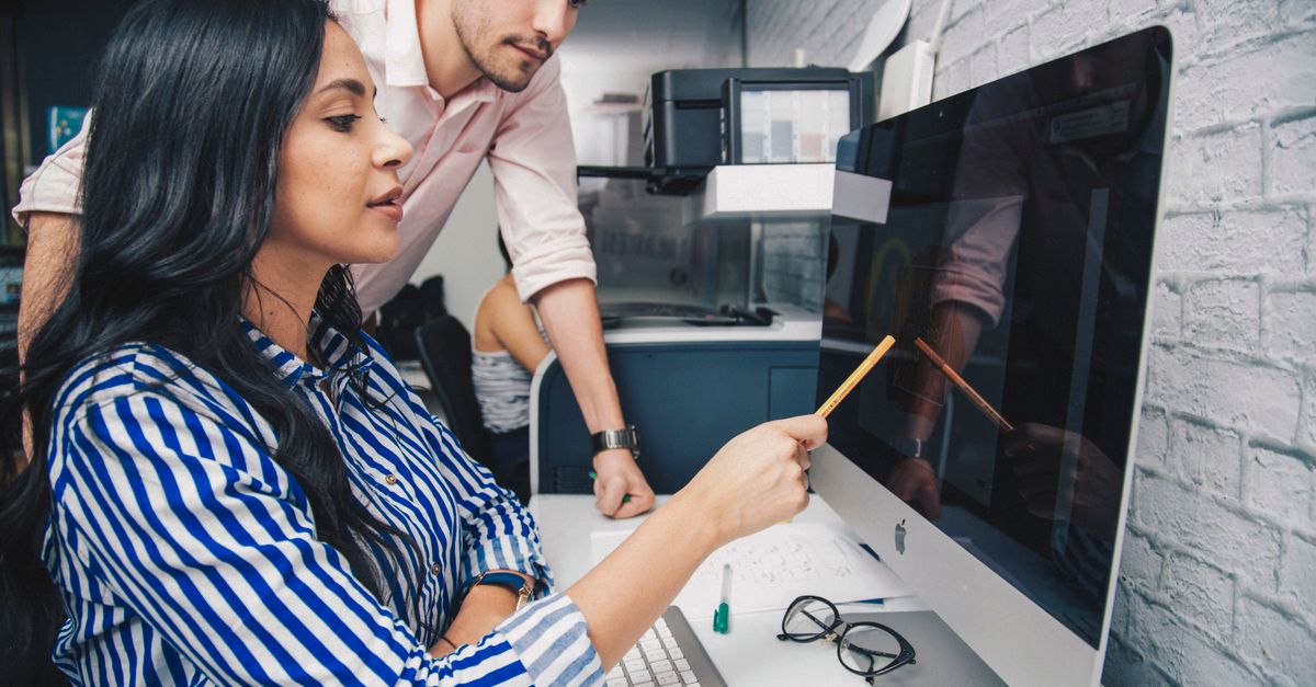 Two coworkers review a desktop screen together, one pointing at it with a pencil while the other leans in.